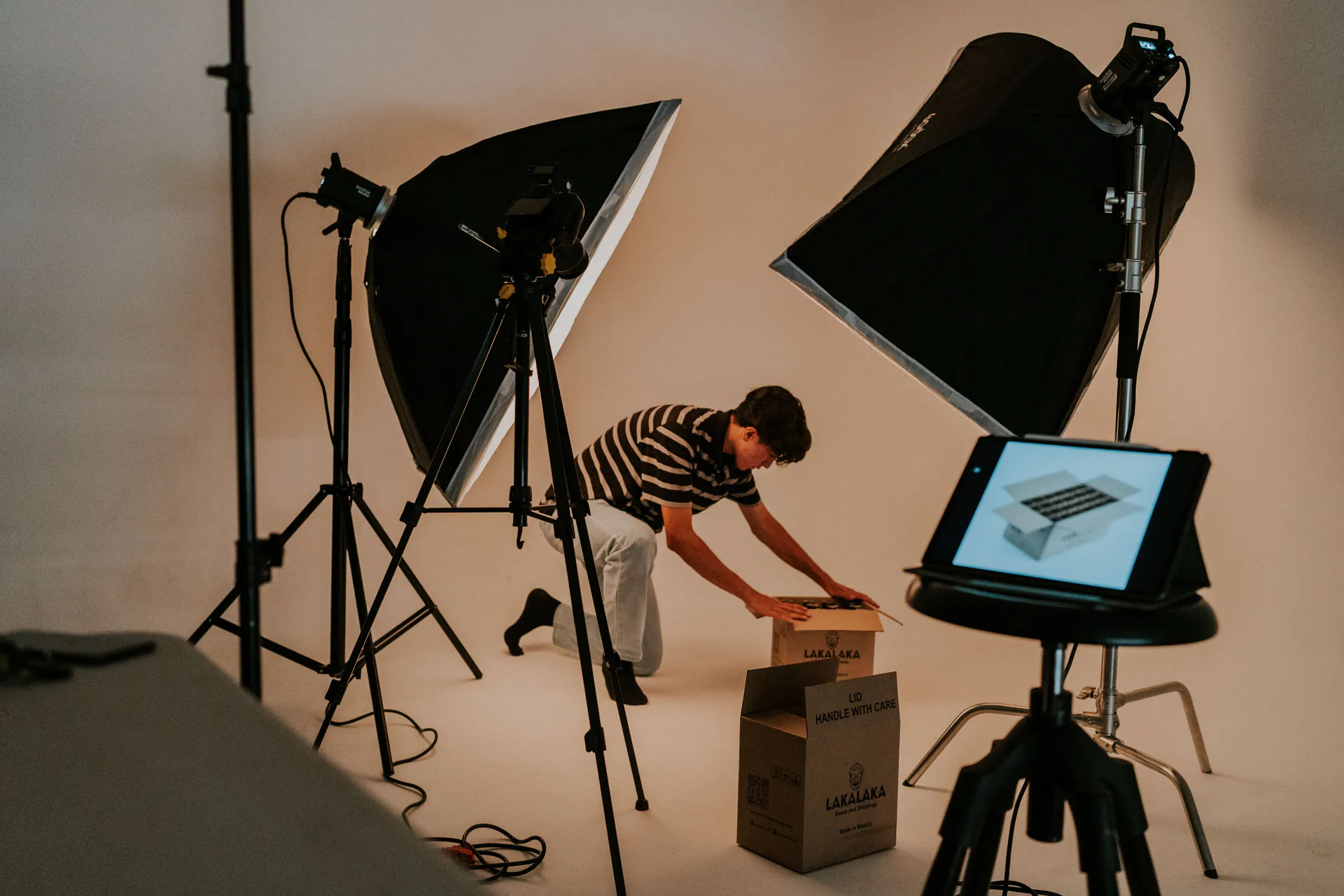 A photographer adjusting a package on a studio table between two softbox lights, with a tablet showing the shot